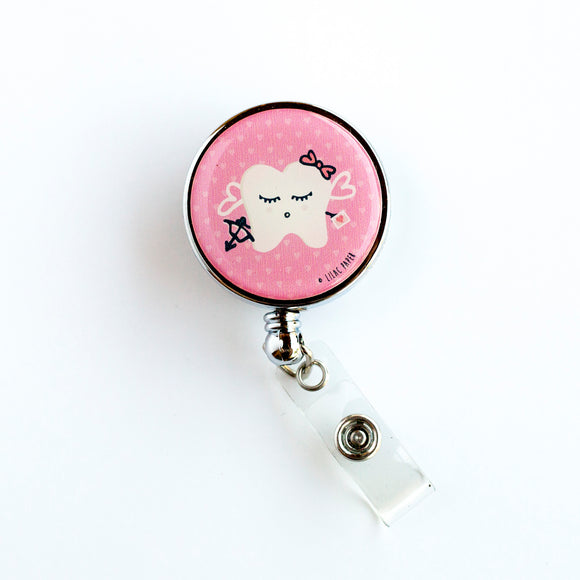 Dental badge reel with white tooth and sweet face dressed up like Cupid complete with bow, arrow, and pink hair bow on a pink heart background. Pink Cupid dental badge reel for dental professionals by Lilac Paper.