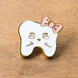 A sweet tooth enamel pin of a smiling tooth with a pink bow. Designed by Lilac Paper.